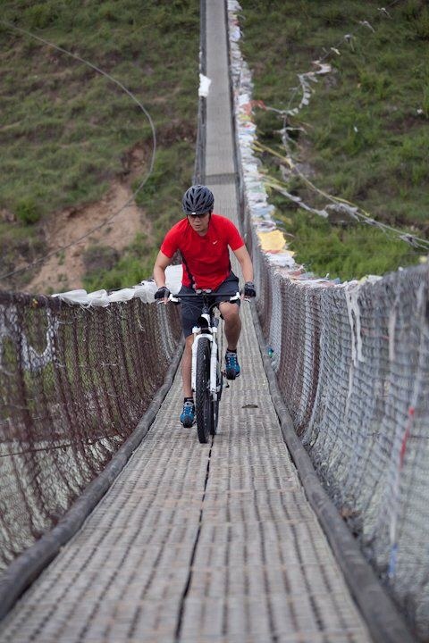A mountain biker in a red jersey riding towards the camera across a narrow suspension bridge strung with prayer flags, a grassy hillside rising behind
